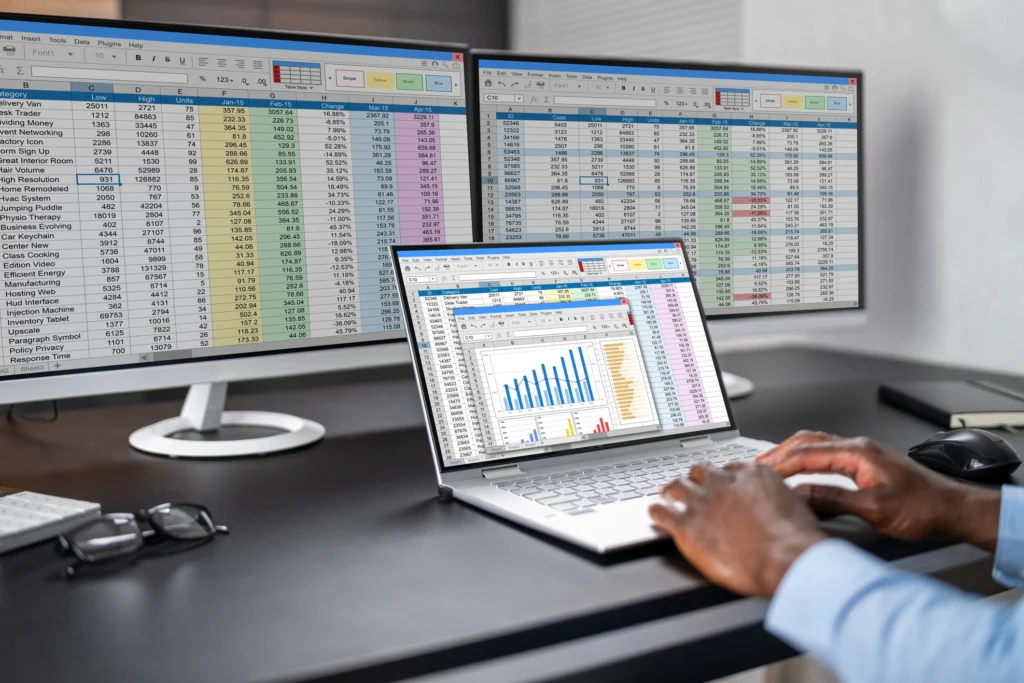 Hände einer Person, die auf der Tastatur eines Laptops tippt, der vor zwei Monitoren steht, auf denen detaillierte Excel-Tabellen geöffnet sind. Der Laptop-Bildschirm zeigt eine Excel-Tabelle mit einem farbigen Balkendiagramm.