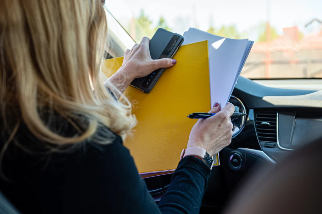 Eine Frau sitzt auf dem Fahrersitz eines Autos, ihr Gesicht ist nicht sichtbar. Sie hält eine gelbe Mappe mit Papieren und ein Handy. Sie sortiert sich gerade, um Ihre vergangene Fahrt in Ihr Fahrtenbuch einzutragen.