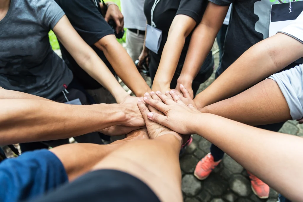 Eine Gruppe von Menschen steht im Kreis und legt ihre Hände übereinander in der Mitte als Zeichen der Einigkeit und Teamarbeit, möglicherweise während eines Treffens zur Gründung eines Vereins. Verschiedene Kleidungsstile deuten auf eine vielfältige Gruppe hin, die sich gemeinsam für ein Ziel engagiert.