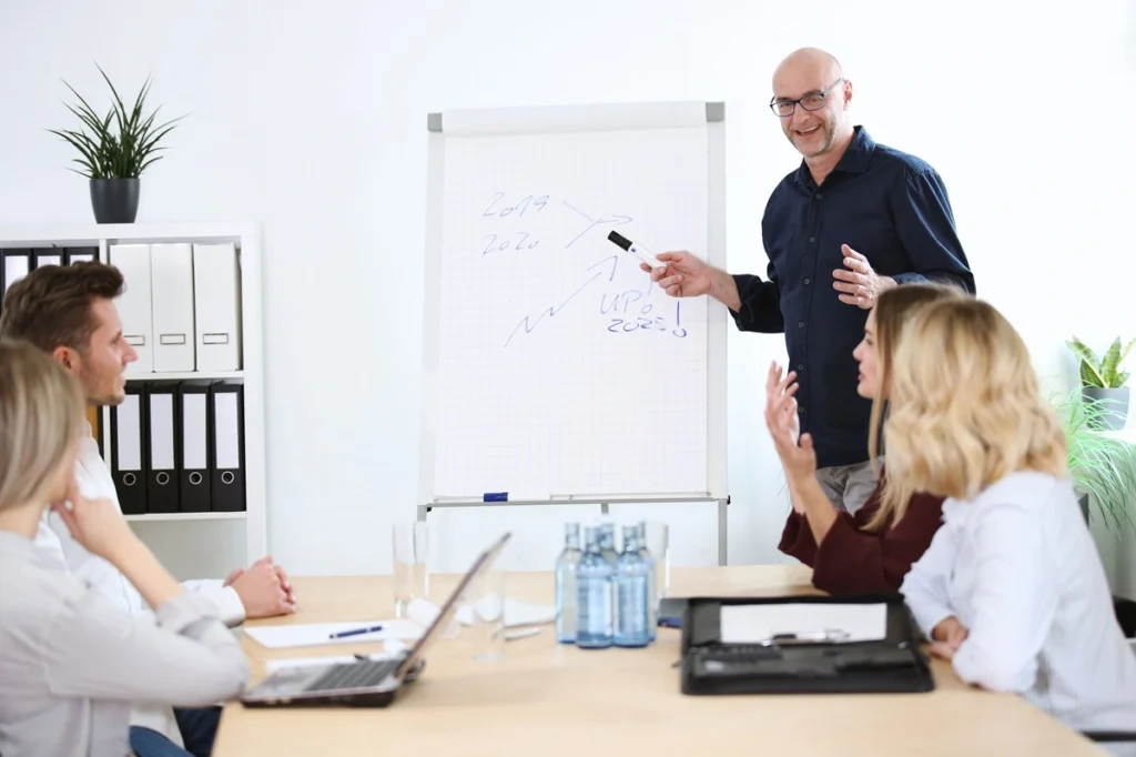 Ein Mann präsentiert auf einem Flipchart. Vier Personen sitzen an einem Tisch und hören zu. Im Kontext der Weiterbildung, werden hier gerade Mitarbeiter weiterqualifiziert.