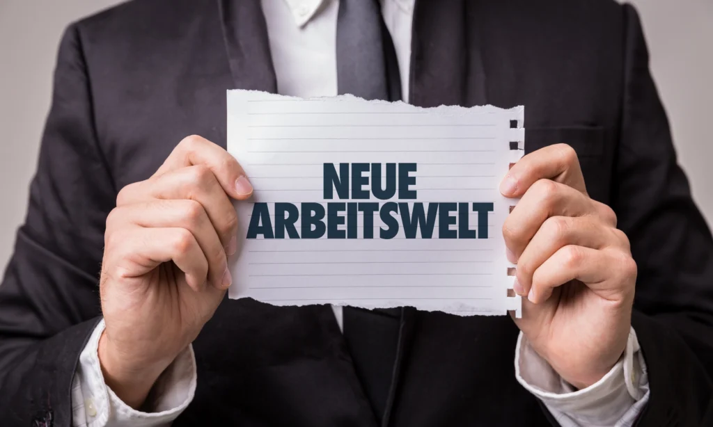 Ein Mann in einem Anzug hält ein zerrissenes Stück Papier vor sich, auf dem in großen Buchstaben 'NEUE ARBEITSWELT' steht. Die zentral platzierte Botschaft auf dem Blatt Papier wird durch die formelle Kleidung des Mannes unterstrichen und deutet auf einen Wandel oder eine neue Ära im Arbeitsleben hin.