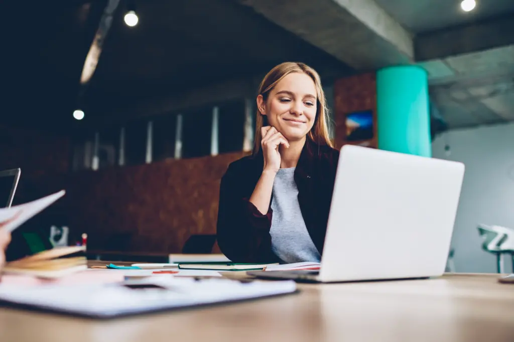 Junge Frau sitzt in modernem Büro am Laptop und lächelt zufrieden beim Arbeiten.