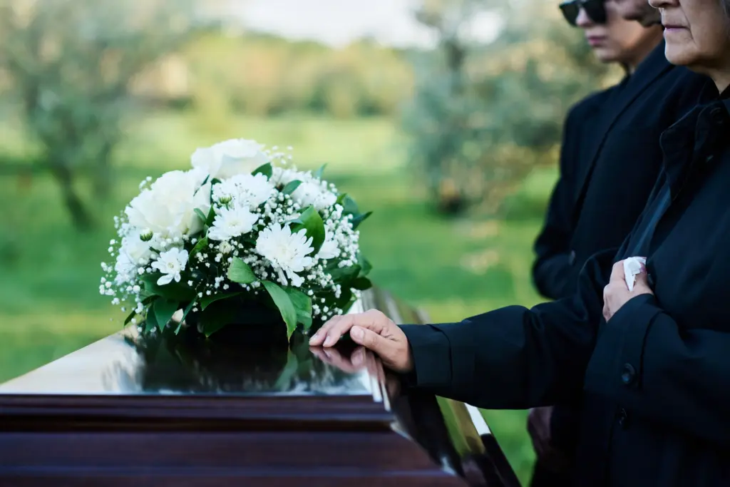 Trauernde in Schwarz stehen an einem Sarg mit Blumenarrangement aus weißen Rosen und Chrysanthemen.